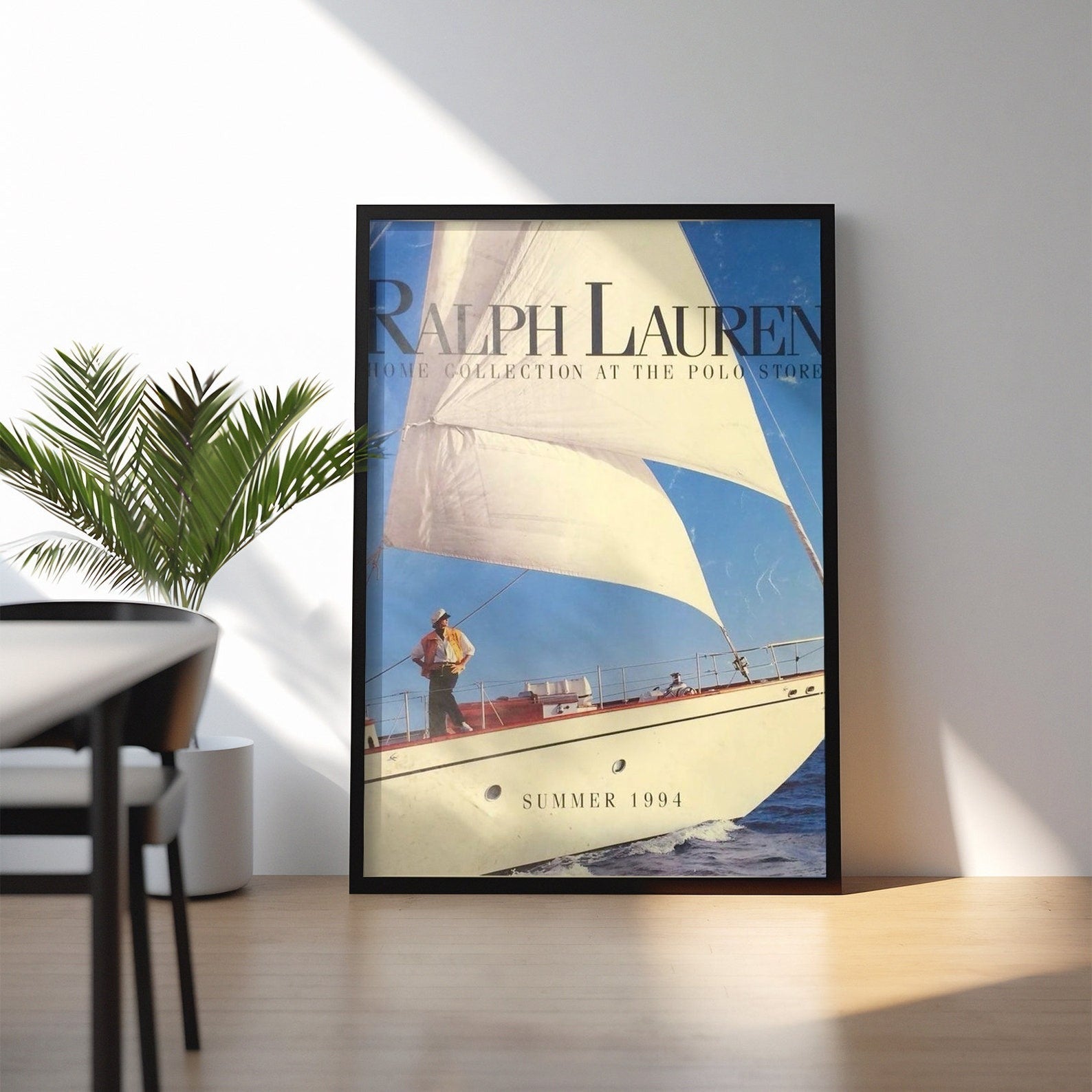 Black framed vintage sailboat poster leaning on a wooden floor next to a green plant in a bright minimalist room with window shadows.