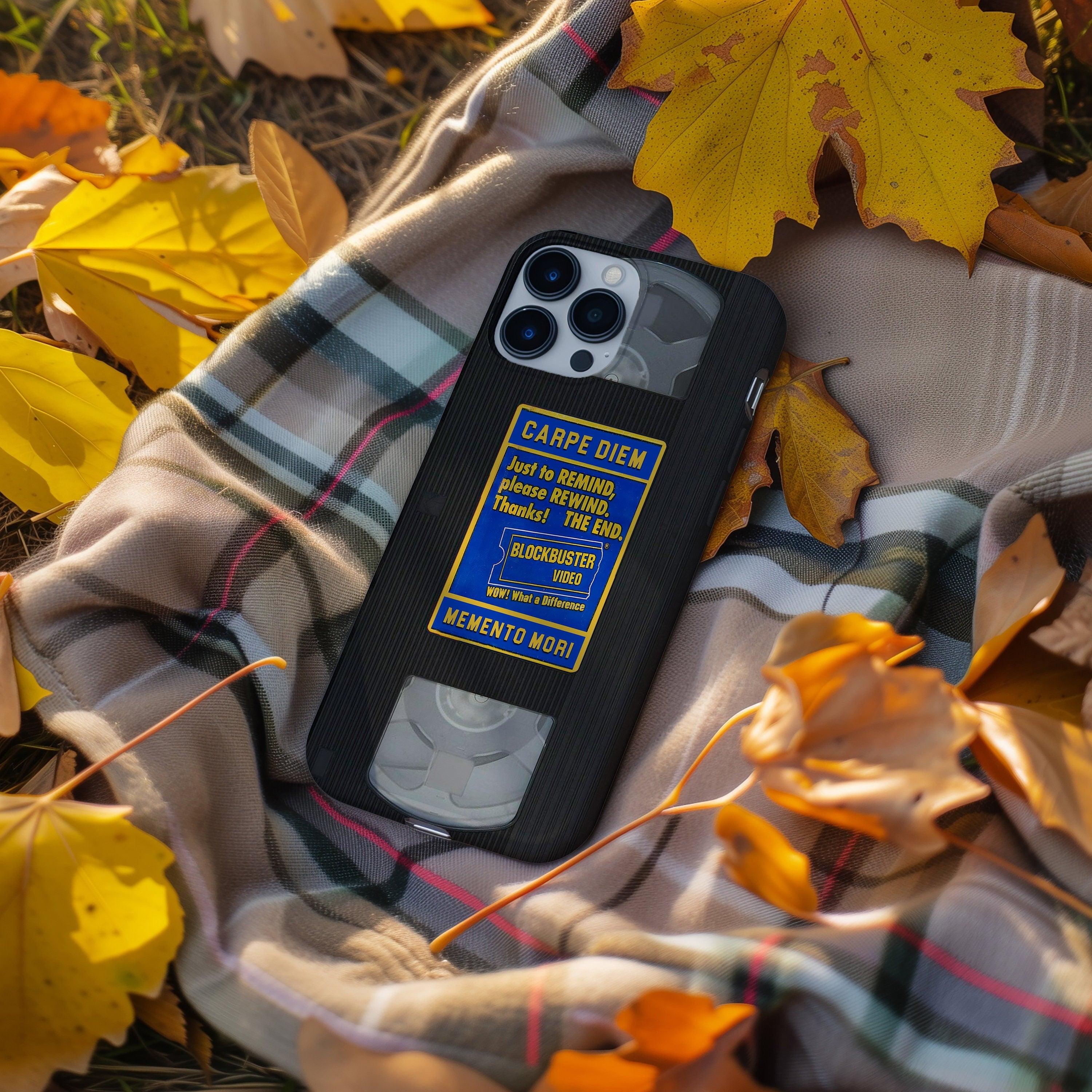 Black VHS cassette iPhone case on plaid blanket among yellow autumn leaves, Memento Mori rental label in view