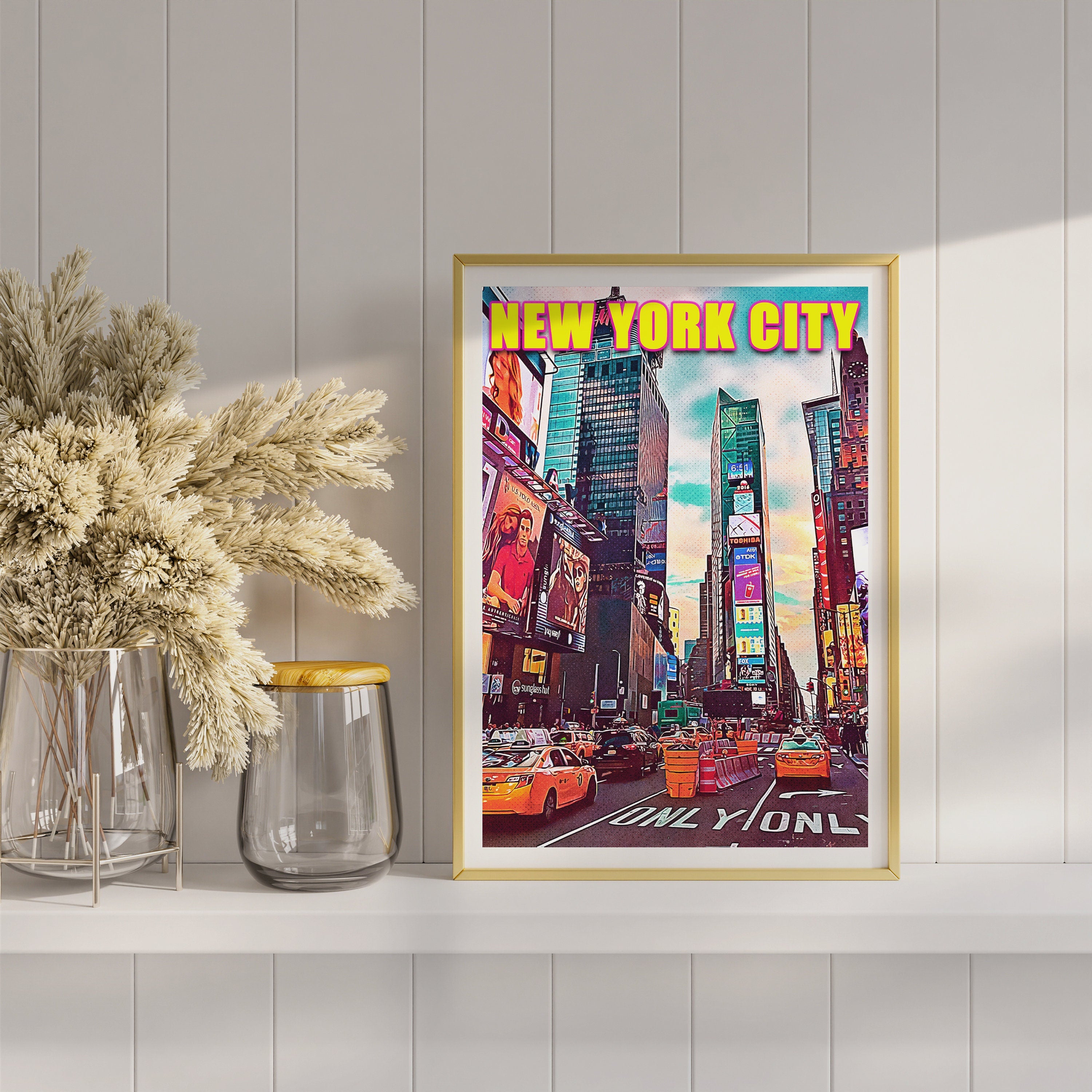 Gold framed Times Square artwork on a white shelf next to dried pampas grass and glass vases